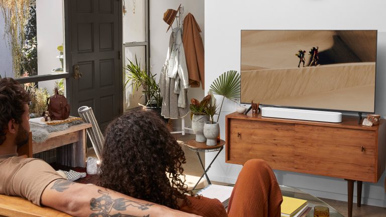 Zwei Personen sitzen im Wohnzimmer auf der Couch und sehen einen Film auf einem Smart TV mit Ton aus einer Soundbar direkt davor.
