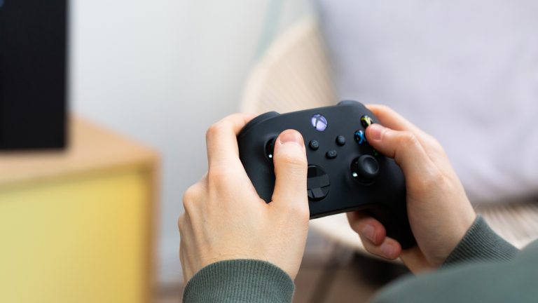 Xbox, Wie du Spiele zurückgeben kannst Eine Person hält den Controller einer Xbox Series X in der Hand. Die Konsole selbst ist unscharf im Hintergrund zu erkennen.