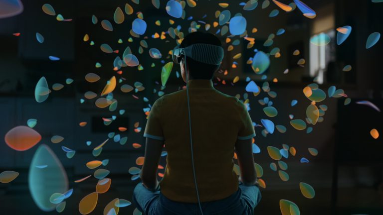 Apple Vision Pro, So richtest du das Headset ein Eine Person mit einer Apple Vision Pro auf dem Kopf sitzt in einem Raum. Um sie herum schweben digiale Inhalte.