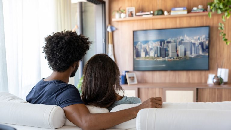Ein Pärchen sitzt im Wohnzimmer auf der Couch und sieht auf einem Fernseher fern. 