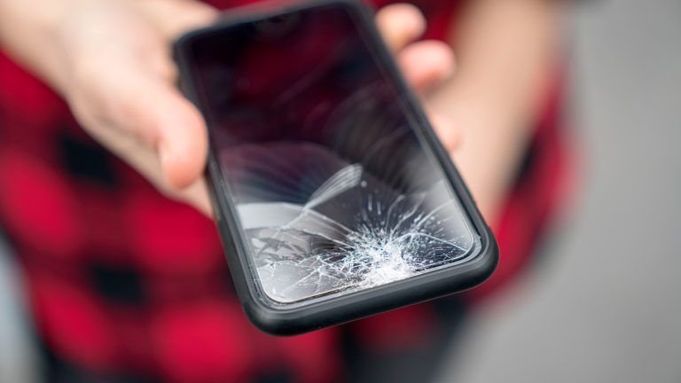 Eine Person hält ein Smartphone in die Kamera, dessen Display gebrochen ist.