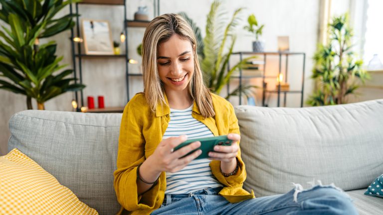 Zattoo, Live-TV, Kosten, Aufnahmen & mehr Eine Person sitzt auf einem Sofa und schaut lächelnd auf einem Smartphone etwas an.