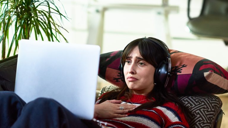 Zattoo, Live-TV, Kosten, Aufnahmen und mehr Eine Person liegt auf einem Sofa, trägt Kopfhörer und schaut etwas auf einem Laptop.