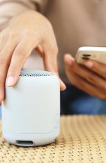 Eine Person hat ein Smartphone in der Hand. Mit der anderen Hand stellt sie einen kleinen Wireless Speaker auf einen Korbtisch.