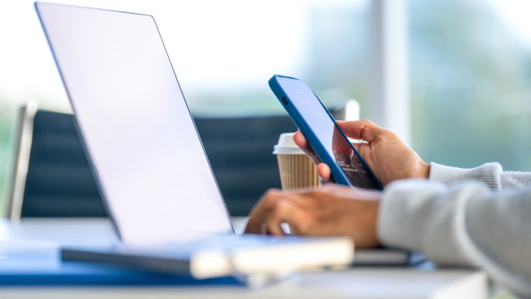 Eine Person hält ein iPhone in der Hand und scheint an einem Schreibtisch zu sitzen.