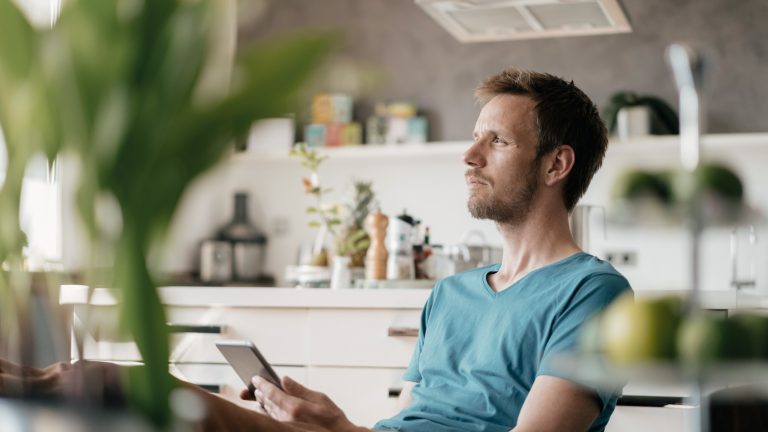 Foodwaste, So vermeidest du es, Lebensmittel wegzuschmeißen Eine Person sitzt am Tisch mit einem Smartphone und blickt nachdenklich in die Ferne.