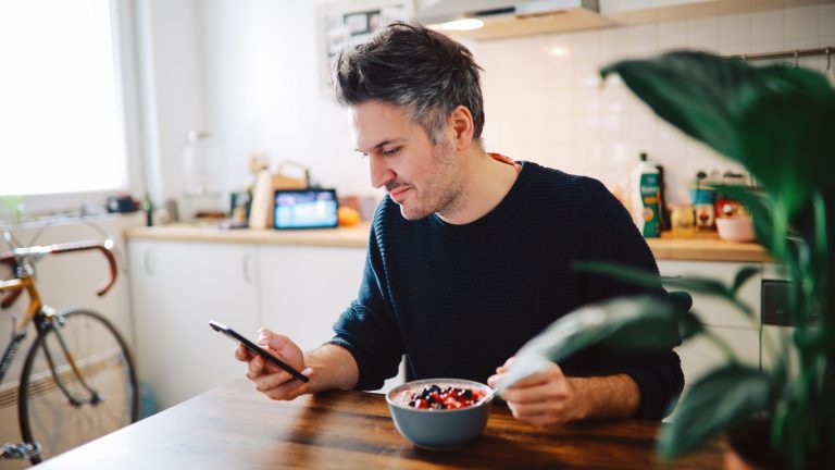 Foodwaste, So vermeidest du es, Lebensmittel wegzuschmeißen Eine Person sitzt am Küchentisch mit einer Schüssel Müsli und schaut ins Smartphone.