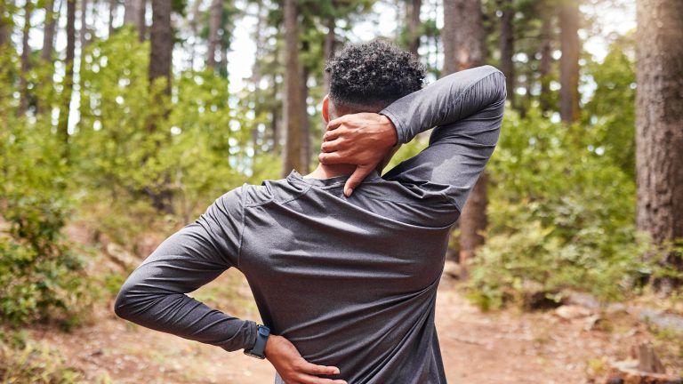 Massagegeräte, Die verschiedenen Typen und Tipps zur Entspannung Eine Person steht in Sportkleidung im Wald und fasst sich in den Nacken.
