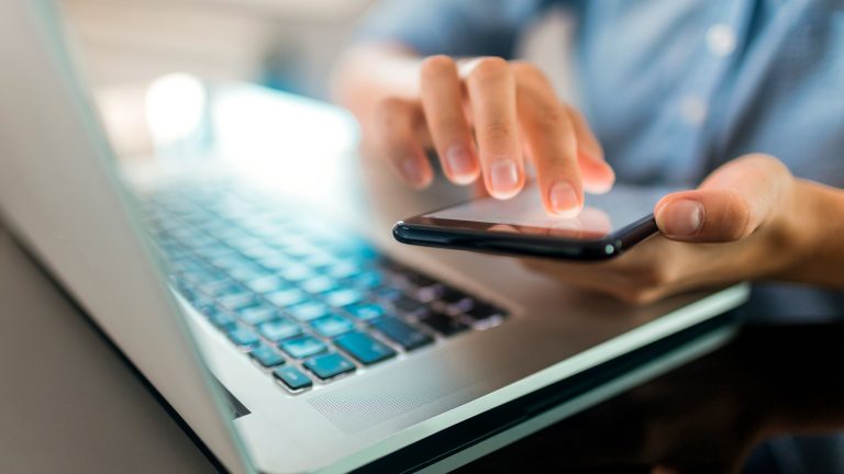 Passkey, Was sie sind und wie sie funktionieren Nahaufnahme eines Laptops und den Händen einer Person, die ein Smartphone bedient.
