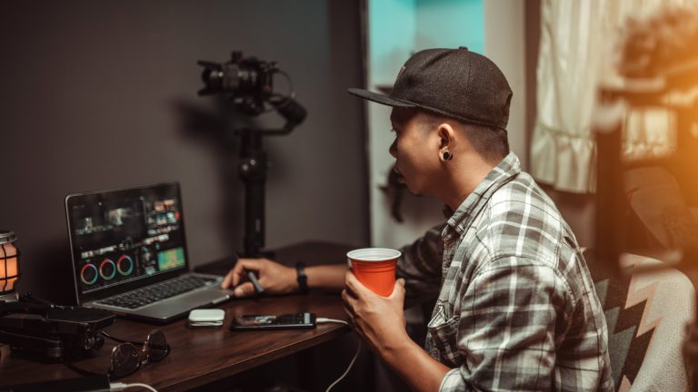 Videos schneiden, Tipps und Anleitungen für dein Video Eine Person mit einem Becher in der Hand sitzt vor einem Laptop, auf dem Videobearbeitung zu sehen ist.