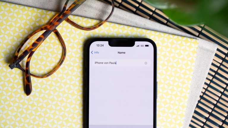 Ein iPhone liegt neben einer Brille auf einem Tisch. Auf dem Display ist das &Auml;ndern des Ger&auml;tenamens zu sehen.