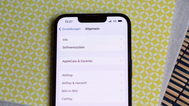 Ein iPhone liegt auf einer gelb-wei&szlig;en Oberfl&auml;che. Auf dem Display sind die Allgemeinen Einstellungen zu sehen
