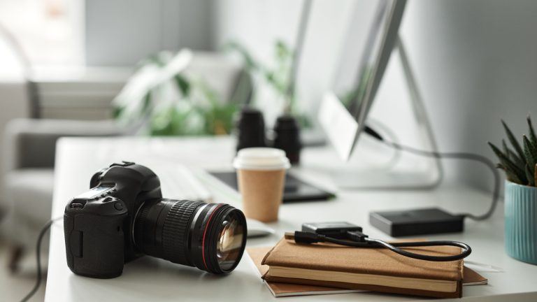 Digitalkamera als Webcam nutzen, So klappt es bei Sony, Canon & Co Eine Digitalkamera liegt auf einem Schreibtisch. Im Hintergrund ist ein Bildschirm und ein Kaffeebecher zu sehen.
