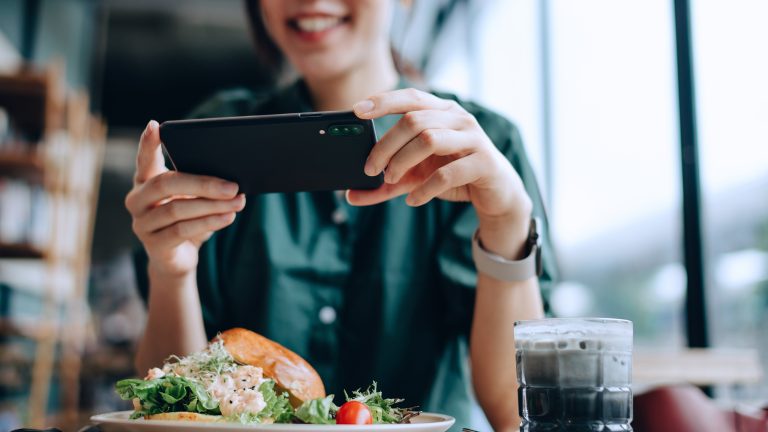 Food Fotografie, Tipps und Tricks um leckeres Essen gut zu inszenieren m Vordergrund steht ein Teller mit Essen, im Hintergrund ist eine Person mit Smartphone, die diesen fotografiert.