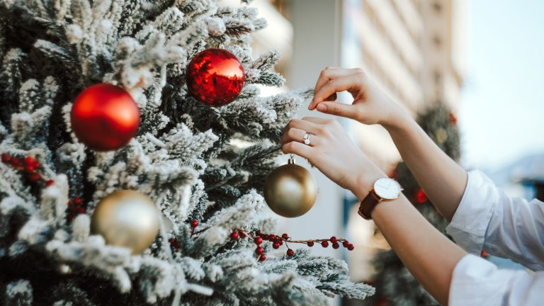Weihnachtsbaeume mit Beleuchtung, die All-in-One-Tanne Nahaufnahme einer All-in-One-Tanne, die mit künstlichem Schnee bedeckt ist. Zwei Hände schmücken diese mit roten und goldenen Kugeln.