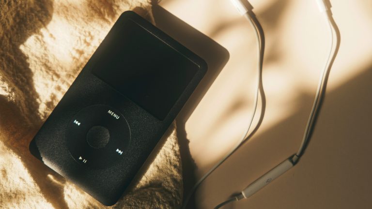 iPod, Warum er auch 2024 noch Sinn macht Ein schwarzer iPod mit weißen Kopfhörern liegt auf einem sandfarbenen Untergrund.