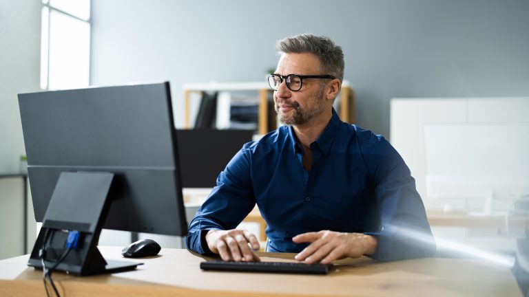 Windows 10 und 11 Taskleiste anpassen, Ausblenden, verschieben und mehr Eine Person von vorn hinter einem großen Bildschirm an einem Schreibtisch in einem cleanen Office.