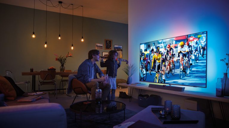 Ambilight, Was ist das und was bedeutet das Plus? Zwei Personen schauen ein Radrennen auf dem Fernseher. Die dahinterliegende Wand ist beleuchet.
