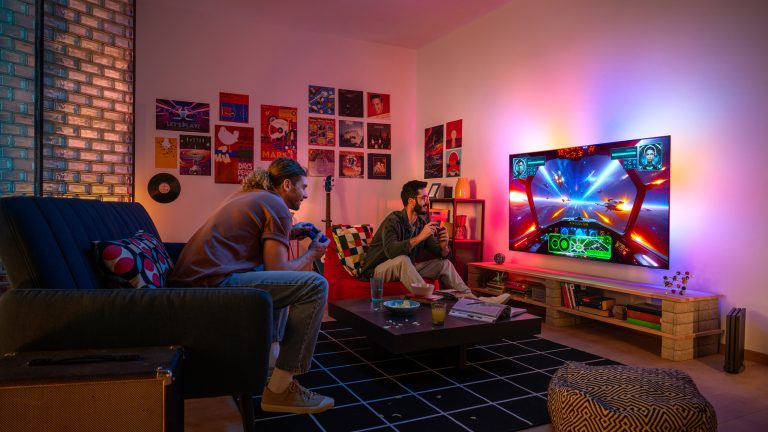 Ambilight, Was ist das und was bedeutet das Plus? Zwei Personen sitzen vor dem Fernseher und spielen ein Videospiel. Die Wand hinter dem Fernseher ist bunt beleuchtet.