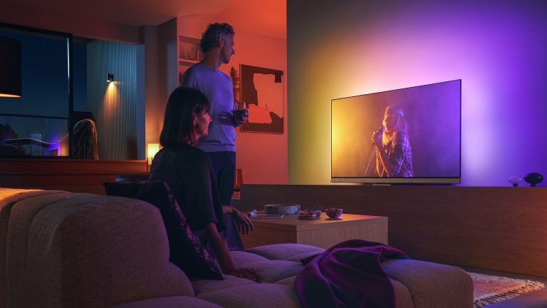 Ambilight, Was ist das und was bedeutet das Plus? Zwei Personen betrachten einen Fernseher in einem Wohnzimmer - eine stehend, eine sitzend. Die Wand hinter dem Fernseher ist durch ihn beleuchtet.