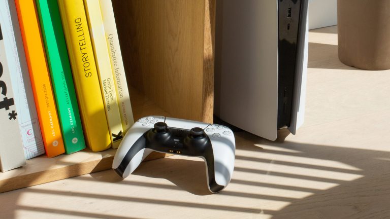 PlayStation Plus, Das kann das Spiele-Abo von Sony Eine PlayStation steht aufrecht in einem Bücherregal mit Büchern und davor liegt ein passender Controller.