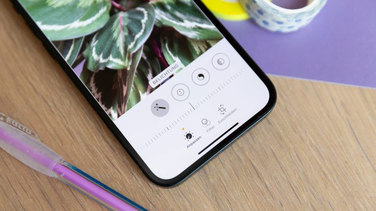 Fotos bearbeiten auf dem iPhone Ein iPhone liegt auf einem Holztisch. Die Fotos-App ist im Bearbeitungsmodus geöffnet und der Regler der Belichtung steht in der Mitte.