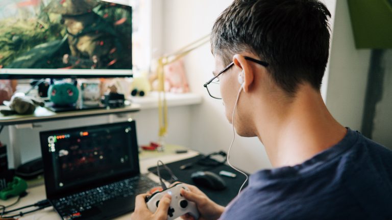 Mini-PC, Was ist das und eignet er sich auch fürs Gaming? Rückansicht einer Person, die vor einem Gaming-Laptop sitzt und einen Controller in beiden Händen hält.