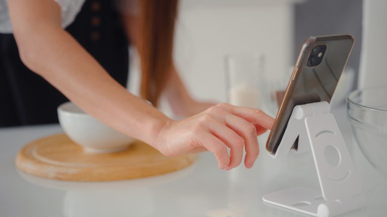 Smartphone, Haltegriffe und Standfüße für mehr Komfort Ein Smartphone steht auf einem Standfuß auf einer Arbeitsfläche in einer Küche. Im Hintergrund ist eine weiße Schüssel auf einem Holzbrett zu sehen.