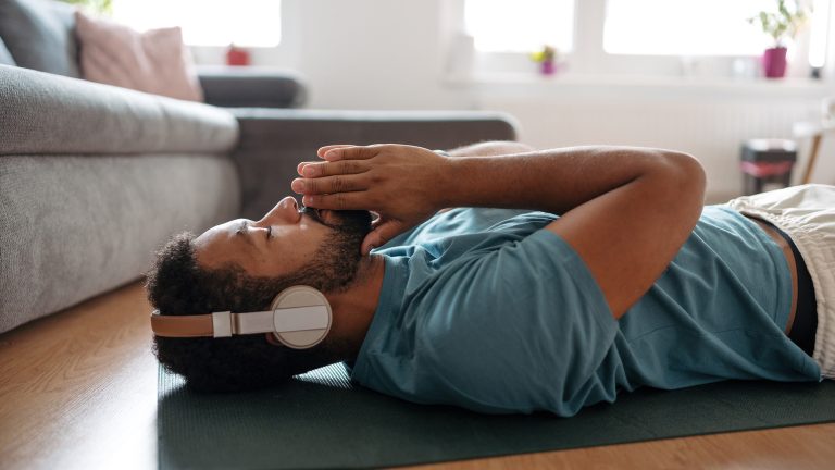 So bringst du mehr Achtsamkeit in dein Leben Eine Person liegt mit geschlossenen Augen auf einer Yogamatte. Sie trägt Over-Ear-Kopfhörer.
