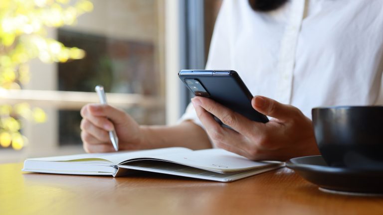 Google Kalender einrichten und nutzen, So geht’s Eine Person sitzt mit einem Stift vor einem Notizblock und hält ihr Smartphone in der anderen Hand.