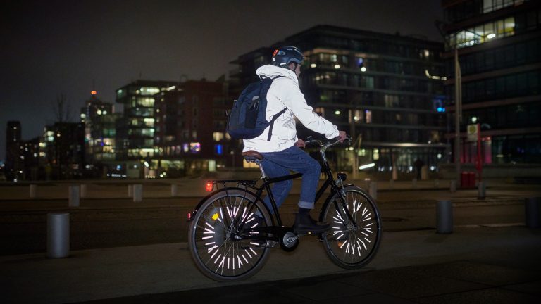 Radfahren, So bist du auch im Winter sicher und mobil Eine Person ist bei Dunkelheit mit einem Fahrrad unterwegs. Jacke und Speichen reflektieren deutlich sichtbar.