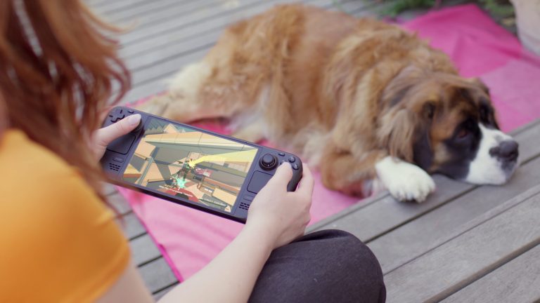 Eine Person sitzt mit einem Steam Deck in der Hand draußen. Im Hintergrund ist ein liegender Hund zu sehen.