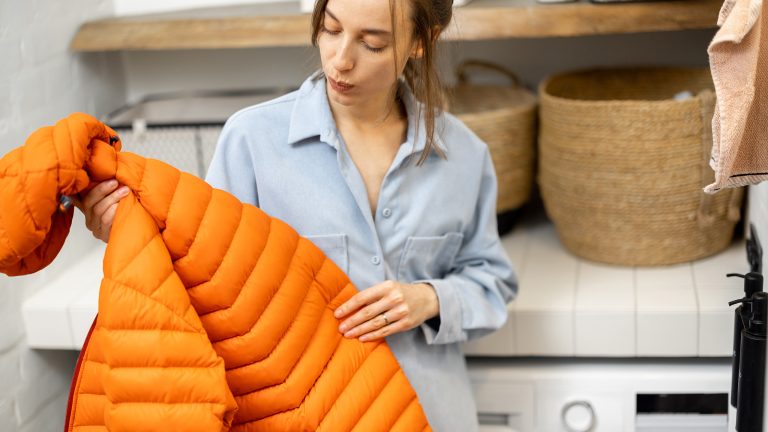 Daunenjacke richtig waschen und trocknen Eine Person steht in einem Waschraum. In der Hand hält sie eine orangene Daunenjacke.