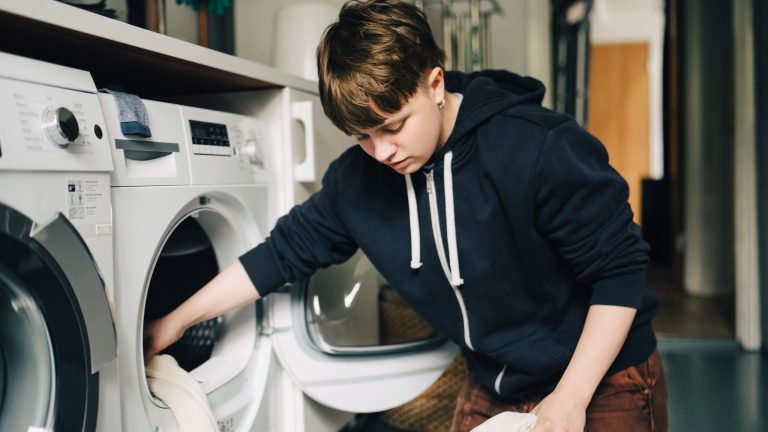 Fleecejacke waschen, Tipps & Tricks für eine gute Pflege Eine Person kniet vor einem Wäschetrockner und einer Waschmaschine und gibt ein Kleidungsstück in den geöffneten Trockner.