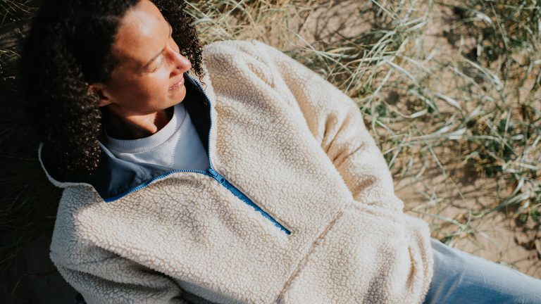 Fleecejacke waschen, Tipps & Tricks für eine gute Pflege Eine Person liegt mit geschlossenen Augen in einem Fleecepullover in einer Sanddüne mit einigen Gräsern.