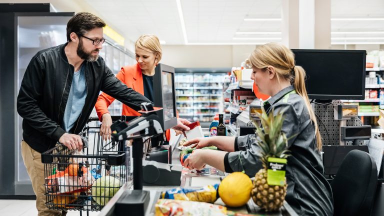 Geld sparen, Diese Apps und Geräte helfen dir dabei Eine Person steht mit einem Einkaufswagen an der Supermarktkasse, Waren liegen bereits auf dem Band. Die Person legt weitere Waren darauf.