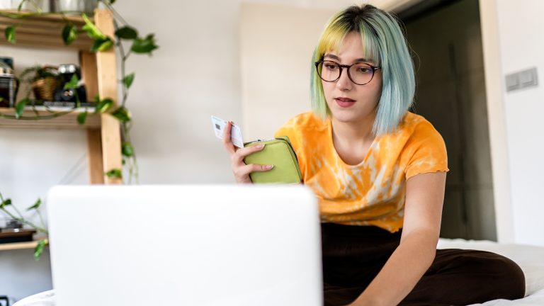 Eine Person sitzt auf einem Bett vor dem geöffneten Laptop. In der Hand hält sie ein Portemonnaie und eine Kreditkarte.