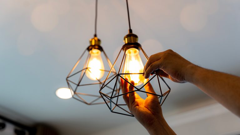 Geld sparen, Diese Apps und Geräte helfen dir dabei Eine Person schraubt eine Glühbirne in eine, an der Decke hängende, Lampe.