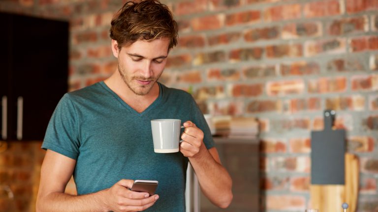 Geld sparen, Diese Apps und Geräte helfen dir dabei Eine Person steht in einer Küche, hält eine Kaffeetasse in der Hand und schaut auf das Smartphone in ihrer Hand.
