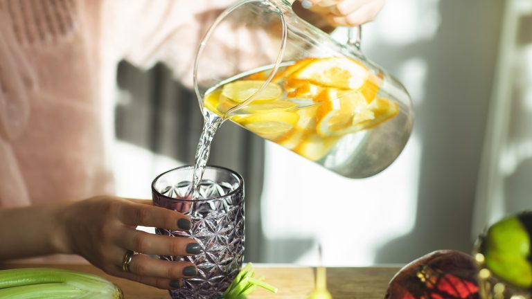 Gesund ernähren leicht gemacht, Mit diesen Geräten klappt’s Eine Person hält ein Glas in der Hand, in das sie Wasser mit Zitronenscheiben aus einer Karaffe gießt.