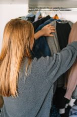 Eine Person steht vor einem Kleiderschrank und schiebt mit beiden Händen Pullover auf Bügeln darin auseinander.