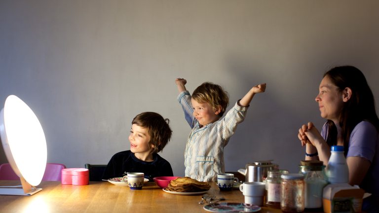 Zwei Kinder und eine erwachsene Person sitzen an einem Tisch und schauen auf eine Tageslichtlampe.