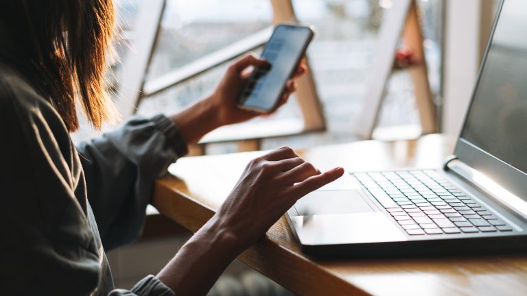 Minimalistisch leben, Mit digitalen Tools und analogen Hilfsmitteln entrümpeln-leben-1 Rückansicht einer Person, die an einem Schreibtisch vor einem Laptop sitzt. In der Hand hält sie auch ein Smartphone.