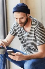 Eine Person sitzt an einem Fenster, hält eine Tasse in der Hand und schaut auf das Smartphone, welches sie in der anderen Hand hält.