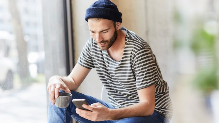 Eine Person sitzt an einem Fenster, hält eine Tasse in der Hand und schaut auf das Smartphone, welches sie in der anderen Hand hält.
