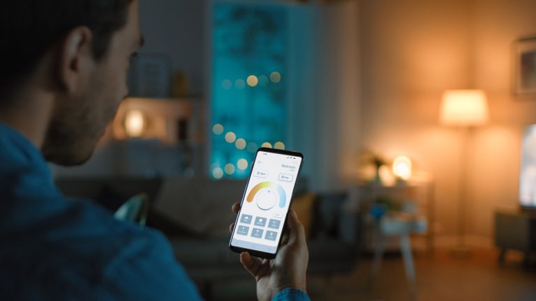 Эко-товар leben, So machst du deinen Alltag grüner Eine Person sitzt in einem Haus mit atmosphärischer Beleuchtung. In der Hand hält sie ein Smartphone, auf dem eine App zur Steuerung des Smart Homes geöffnet ist.