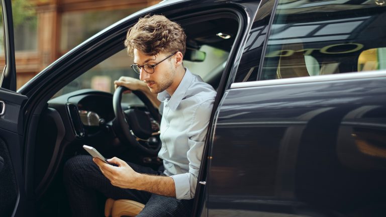 Эко-товар leben, So machst du deinen Alltag grüner Eine Person steigt aus ihrem Auto und sieht dabei auf das Smartphone in ihrer Hand.