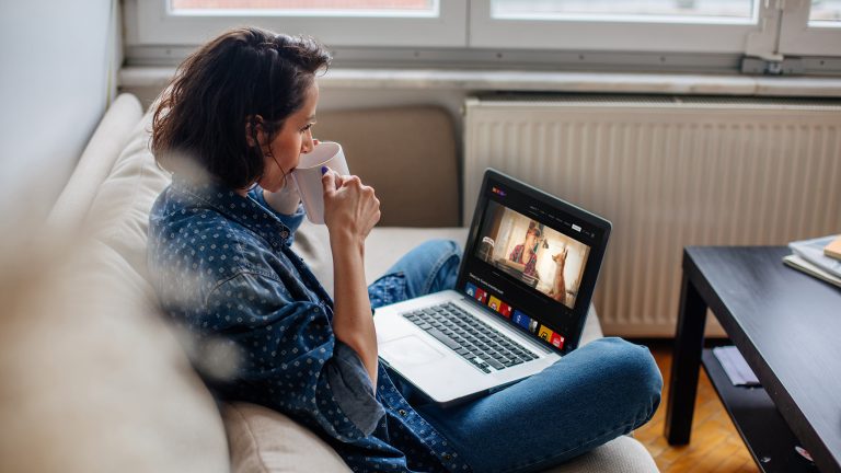 Eine Person sitzt mit einer Tasse auf dem Sofa vor einem Laptop und nutzt RTL+.