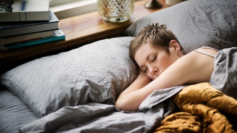 Süße Technik-Träume, Besser schlafen mit diesen Gadgets Eine Person liegt in einem Bett mit grauem Bezug und schläft, während eine Hand unter dem Kopf liegt.