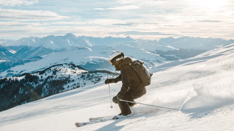 Eine Person fährt auf Skiern die Piste herunter. Im Hintergrund sind schneebedeckte Berge zu sehen.